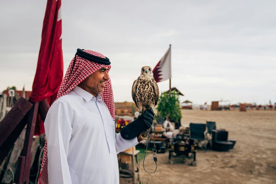 Un uomo in abiti tradizionali arabi tiene un falco sul guanto in un accampamento desertico all'aperto con bandiere.