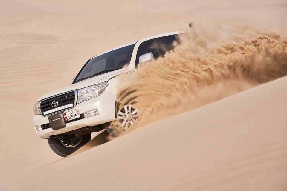 Un SUV bianco guida lateralmente lungo una ripida duna di sabbia, sollevando un'ampia colonna di sabbia nel deserto.