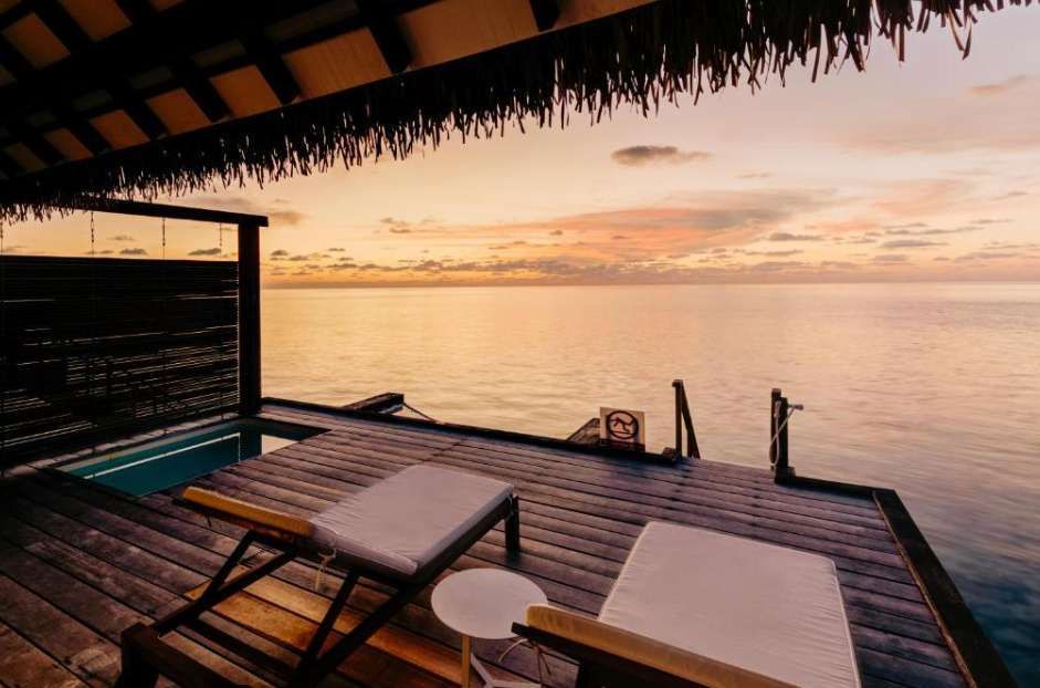 Two lounge chairs and a small pool on a wooden deck of an overwater villa, overlooking a calm ocean at sunset.
