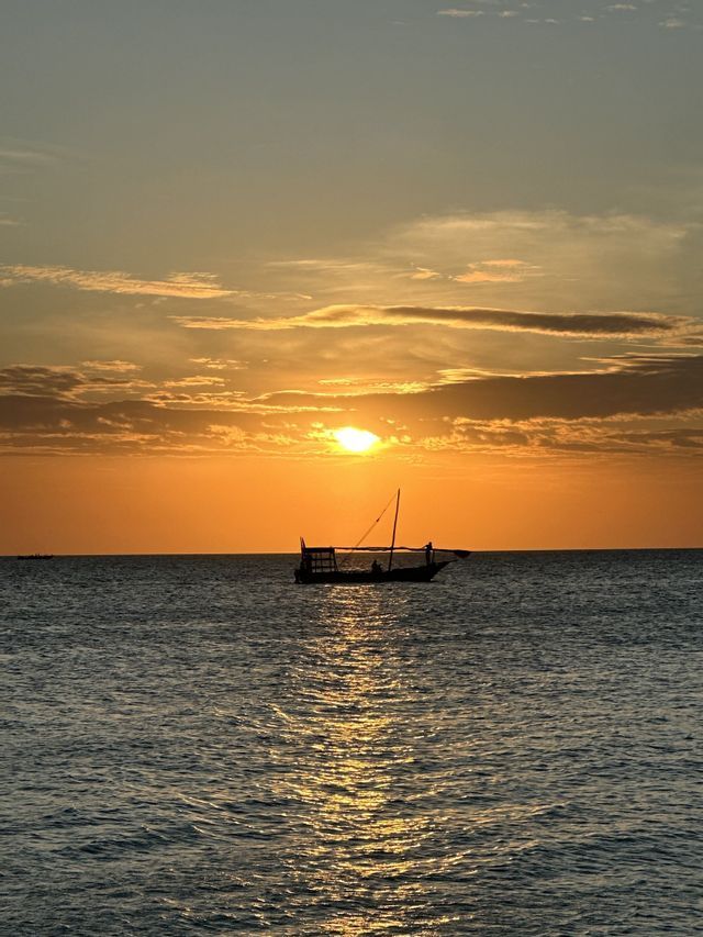 Ein traditionelles Boot als Silhouette gleitet bei einem orangefarbenen Sonnenuntergang auf dem Meer, wobei sich das Licht auf dem Wasser spiegelt.