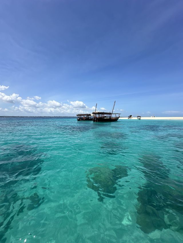 Zwei Holzboote ankern im seichten türkisfarbenen Wasser neben einer weißen Sandbank unter klarem blauem Himmel.