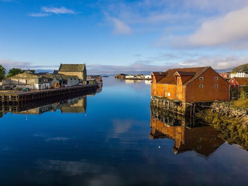 Casas de botes de madera sobre pilotes bordean una ensenada tranquila, sus reflejos espejados en el agua azul inmóvil bajo un cielo despejado.
