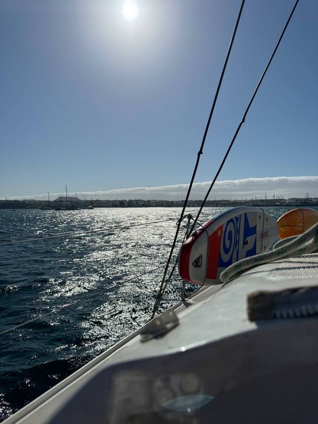 Una tabla de paddle está atada a un velero en aguas bañadas por el sol, con una costa lejana bajo un sol brillante.