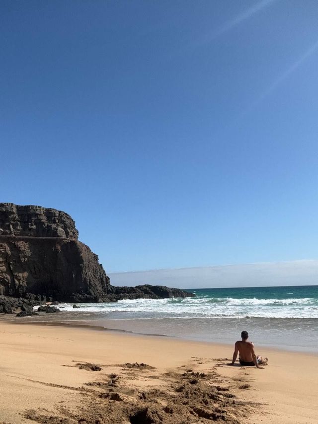 Una persona siede su una spiaggia sabbiosa di fronte all'oceano, con una grande scogliera rocciosa alla sinistra sotto un cielo sereno.