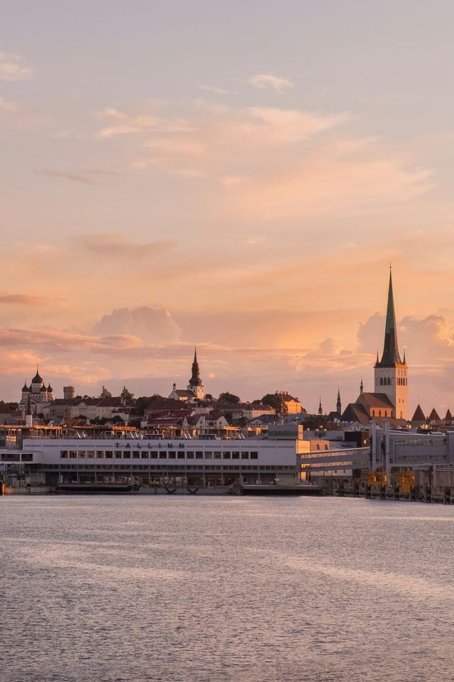 Una vista lungomare dello skyline di una città con guglie storiche e un grande terminal portuale sotto un cielo al tramonto.