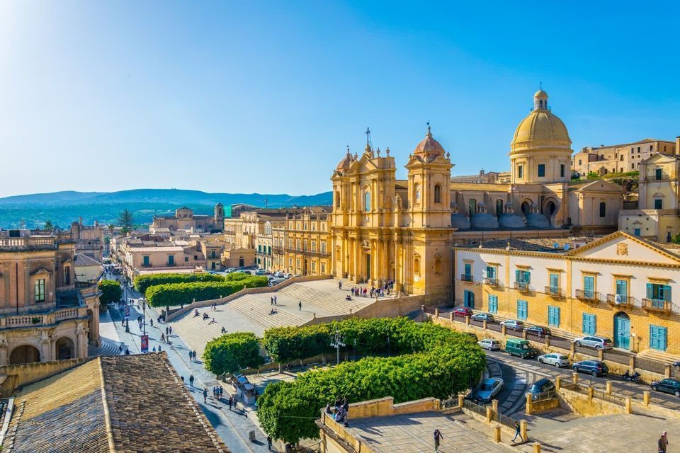 An elevated view of a historic city with a baroque cathedral and a grand staircase leading to a square with people walking.