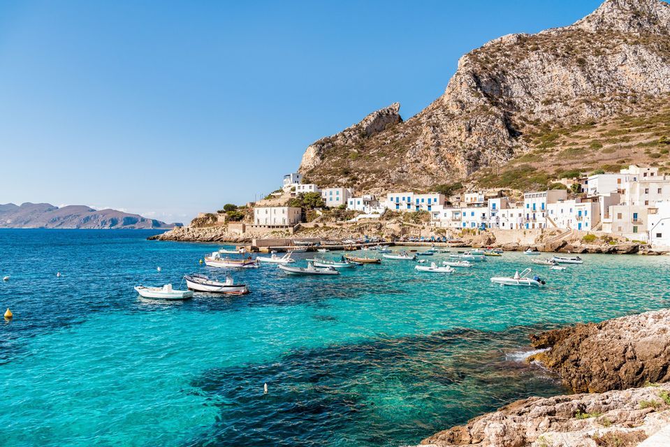 Small boats anchored in a turquoise bay in front of a coastal village with white houses built into the side of a rocky mountain.