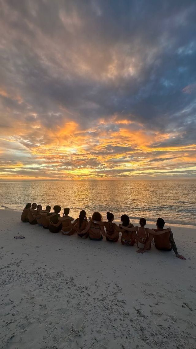 Un grupo de viaje de WeRoad se sienta junto en una playa de arena, observando una colorida puesta de sol sobre el océano.
