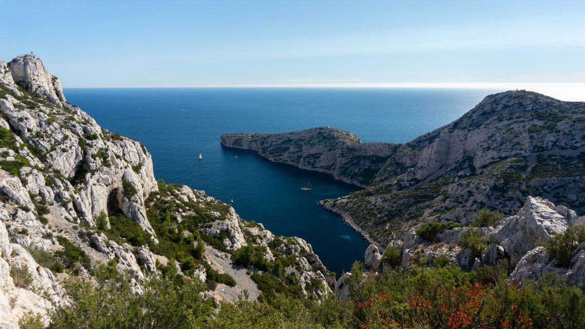 Una vista dall'alto di un'insenatura costiera rocciosa con acqua blu profonda, dove diverse barche a vela sono visibili sotto un cielo limpido.