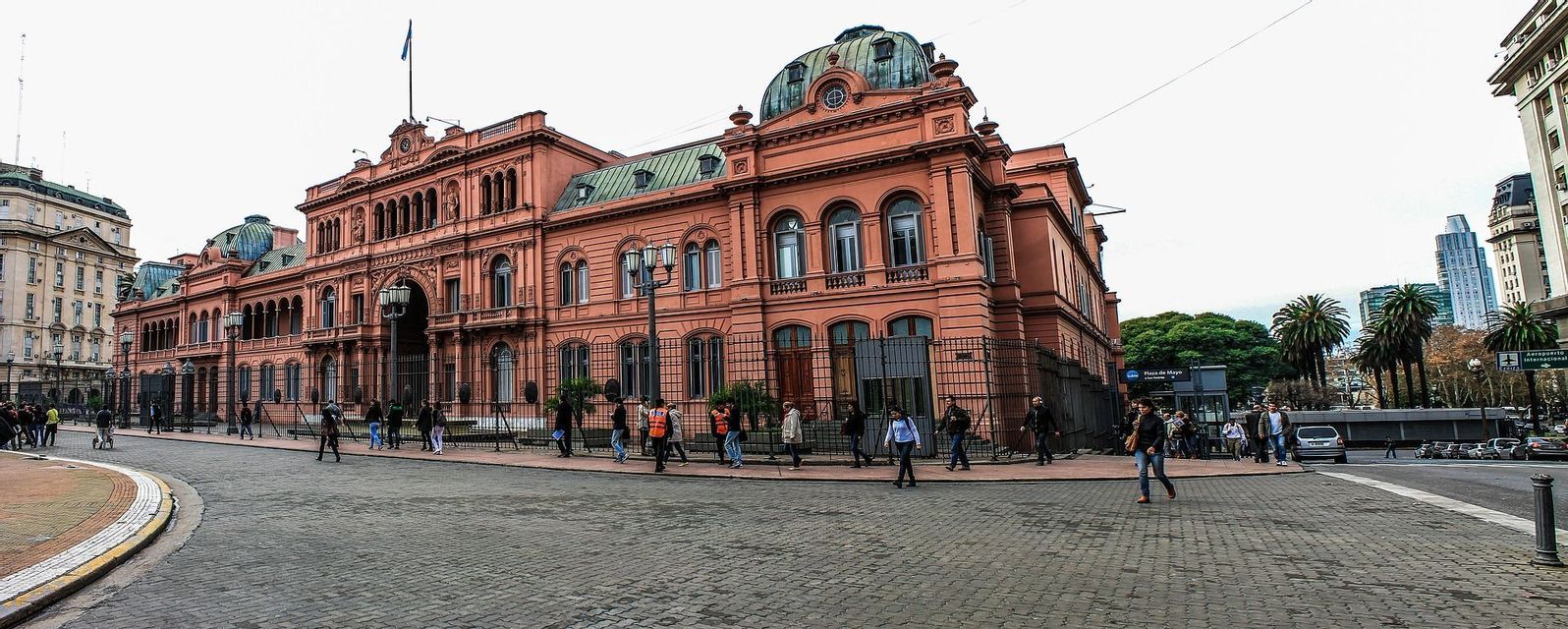 Una vista grandangolare di un edificio governativo rosa e ornato con persone che camminano sulla strada acciottolata di fronte.