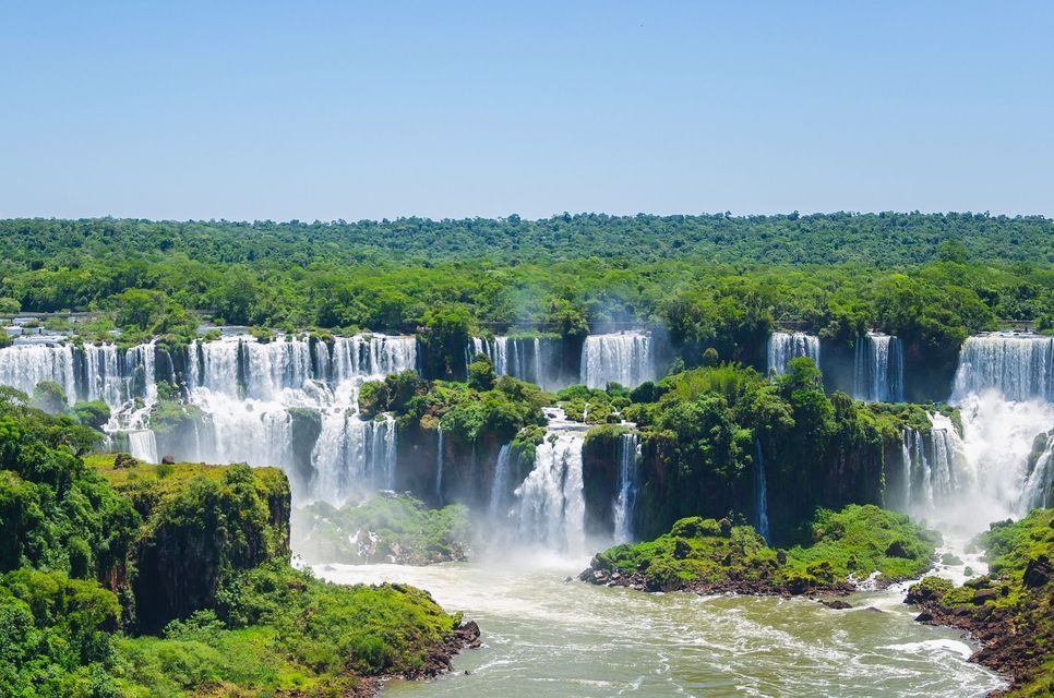 Una veduta panoramica di un vasto sistema di cascate che si riversano su scogliere rocciose ricoperte da una lussureggiante vegetazione verde sotto un cielo blu limpido.