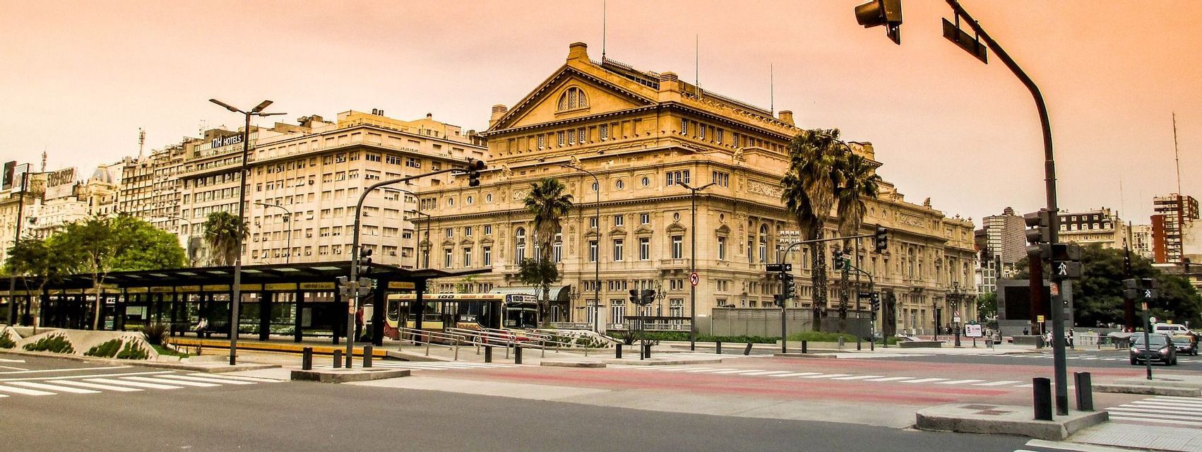 Un edificio classico e ornato all'angolo di una strada cittadina, con un ampio viale, attraversamenti pedonali e una moderna fermata dell'autobus sotto un cielo dalle sfumature arancioni.