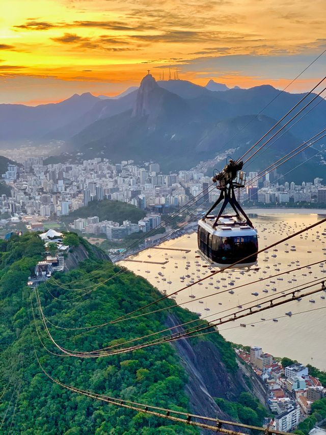 Una funivia viaggia sopra una città costiera e una baia piena di barche, con una grande statua su una cima di montagna al tramonto.