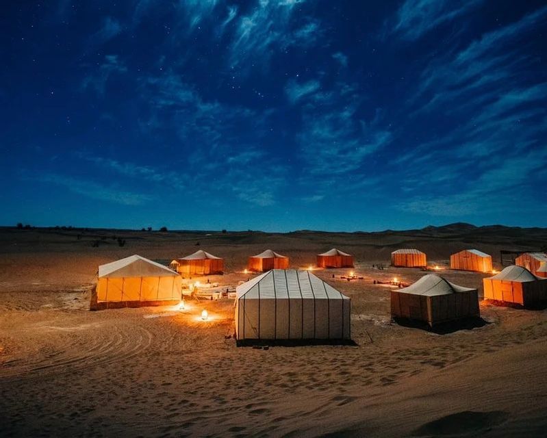 Tende illuminate disposte in un campeggio su dune di sabbia nel deserto sotto un cielo stellato.