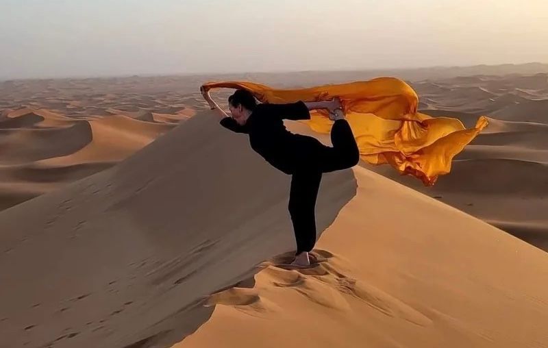 Una donna in nero esegue una posa yoga del ballerino su una duna di sabbia, con un grande tessuto giallo che ondeggia dietro di lei.