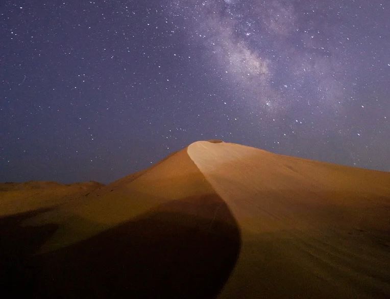 Una grande duna di sabbia nel deserto di notte, con la Via Lattea visibile nel cielo stellato.