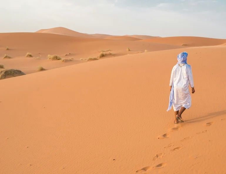 Una persona con una tunica bianca e un turbante blu cammina su dune di sabbia arancione, lasciandosi dietro delle impronte.