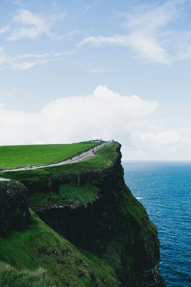 Un viaggio di gruppo WeRoad percorre un sentiero in cima a ripide scogliere verdi a picco sull'oceano, sotto un cielo nuvoloso.