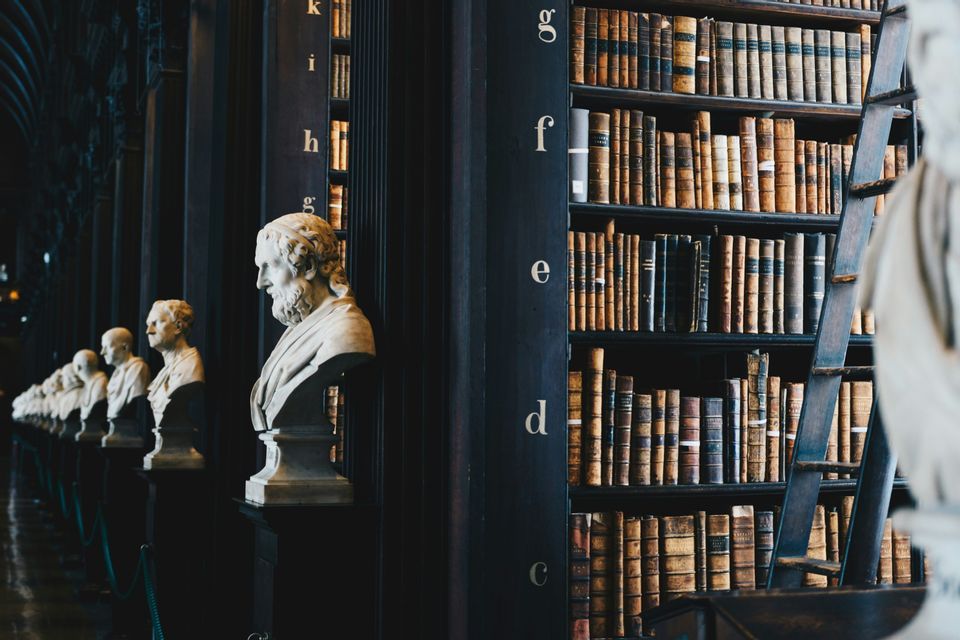 Busti di marmo fiancheggiano una sala tra alte librerie piene di libri antichi in una biblioteca storica.
