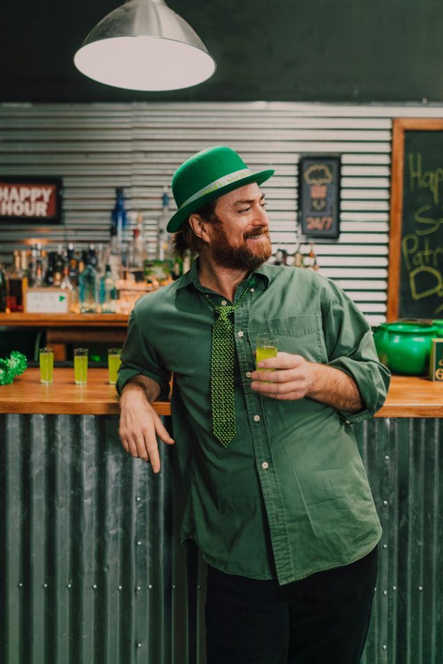 Un uomo con una camicia verde, un cappello e una cravatta a perline tiene un bicchierino appoggiato al bancone di un bar durante i festeggiamenti di San Patrizio.