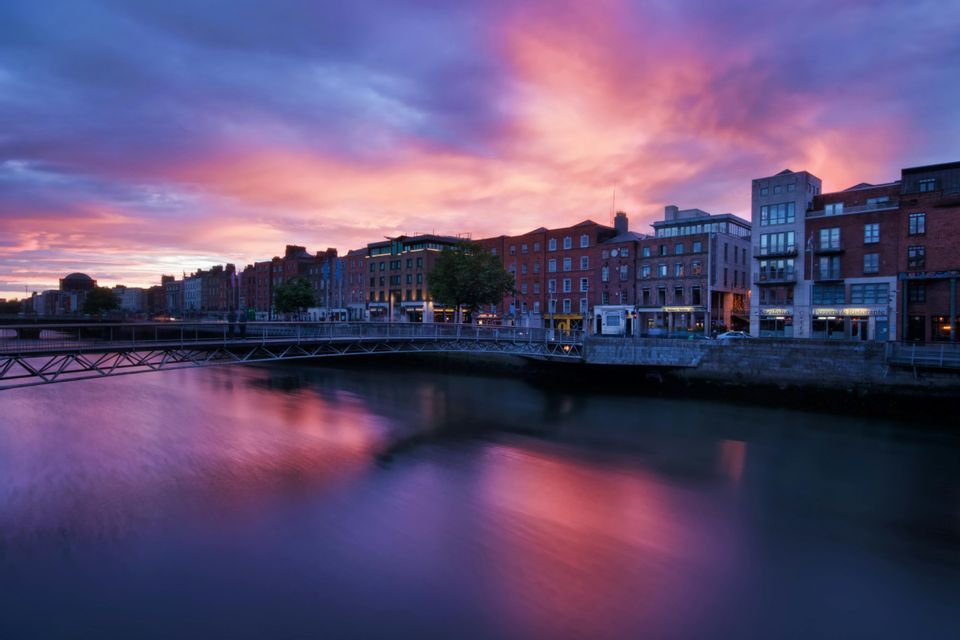 Un fiume riflette i colori viola e rosa di un cielo al tramonto mentre scorre tra gli edifici della città e sotto un ponte pedonale.