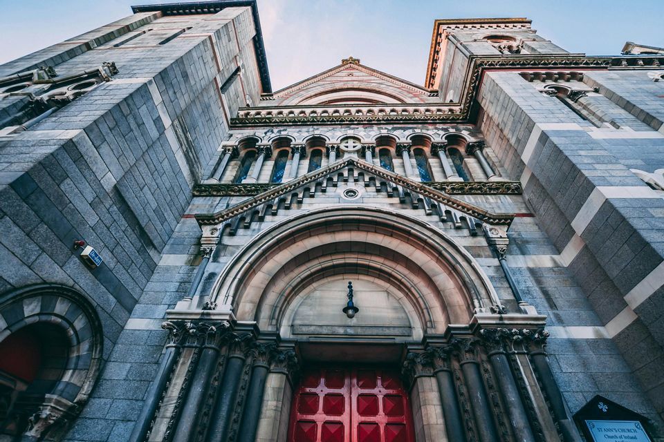 Una vista dal basso della facciata in pietra ornata di una grande chiesa con torri gemelle e una prominente porta rossa.