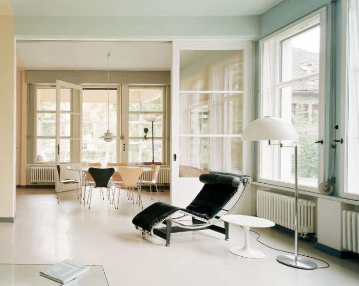 Un soggiorno moderno e luminoso con una chaise longue nera, una lampada da terra bianca e una zona pranzo visibile attraverso un'ampia apertura.