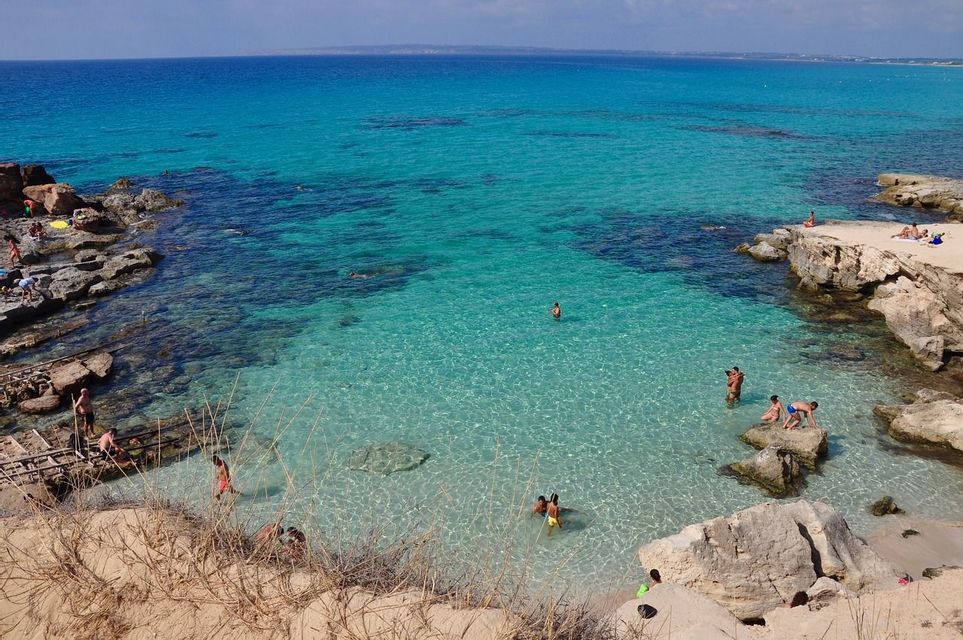 People swimming and sunbathing in a rocky cove with clear, turquoise water under a sunny sky.