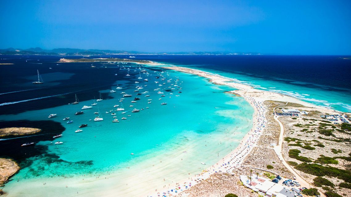 An aerial view of a narrow sand spit with boats anchored in the turquoise water on one side and the deep blue sea on the other.