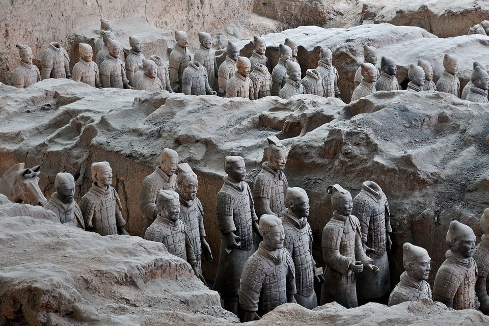 Estatuas de guerreros de terracota de tamaño natural se encuentran en formación en una fosa de excavación arqueológica, parcialmente cubiertas por tierra.