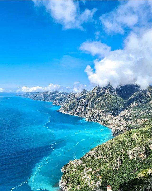 Vista dall'alto di una costa montuosa e verdeggiante che si affaccia su un mare turchese e blu vibrante sotto un cielo parzialmente nuvoloso.