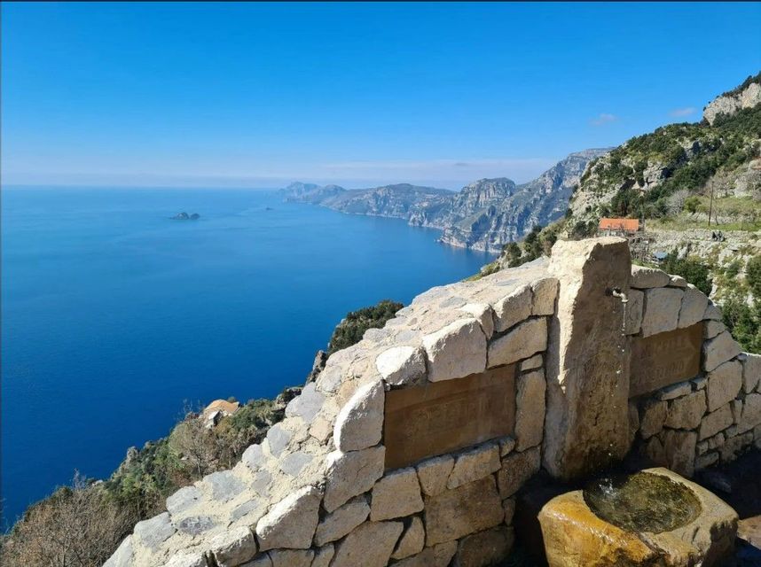 Una fontana in pietra su una scogliera si affaccia su una costa frastagliata e un mare blu profondo sotto un cielo sereno