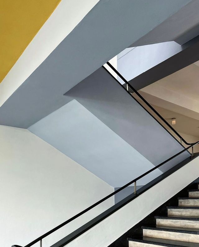 Una vista astratta, dal basso, di una scala con pareti geometriche in bianco, grigio e giallo, con un corrimano nero.