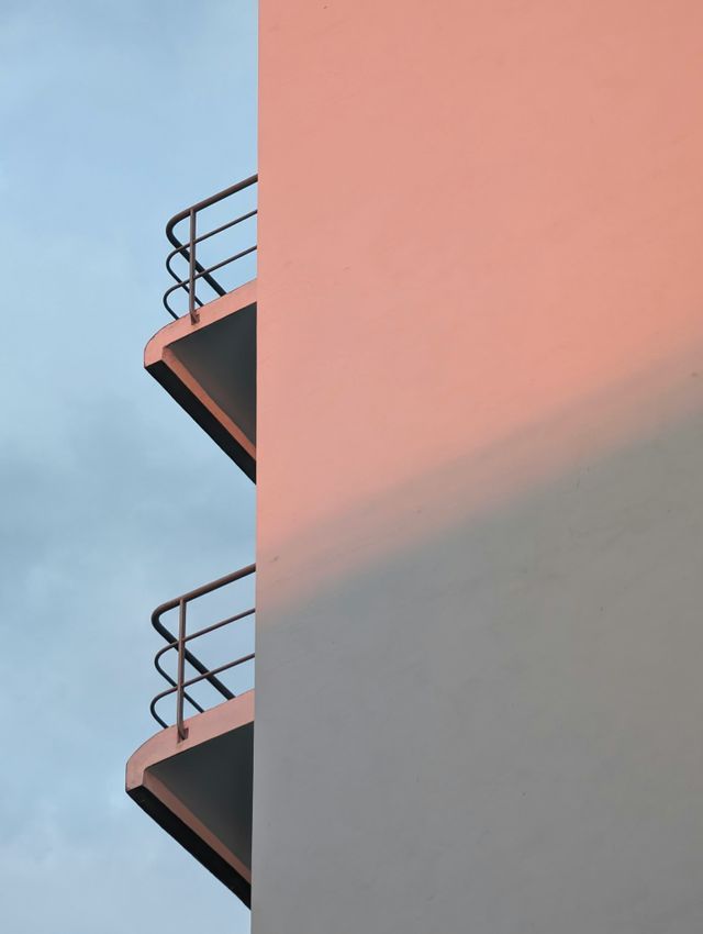 Scatto minimalista: angolo di edificio rosa con due balconi e ombra netta su cielo blu