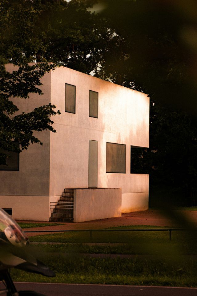 Un edificio bianco e modernista con finestre geometriche si erge tra alberi scuri, la sua facciata illuminata da luce calda.