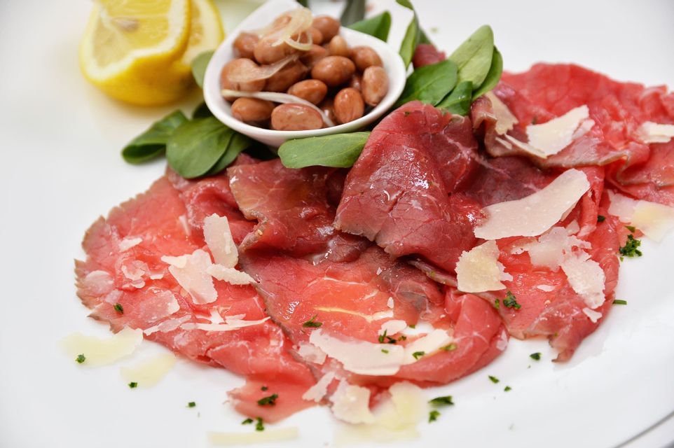 Carpaccio di manzo con scaglie di parmigiano e verdure su un piatto bianco, accanto a una ciotola di fagioli e spicchi di limone fresco.