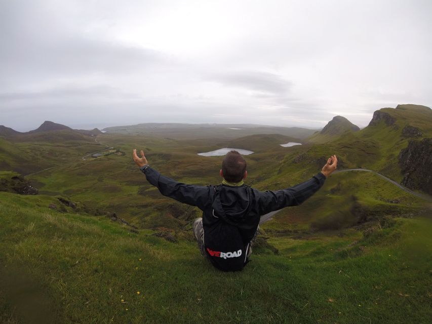 Une personne portant un sac à dos WeRoad est assise sur une colline verdoyante, les bras ouverts, contemplant une vallée parsemée de lacs sous un ciel nuageux.