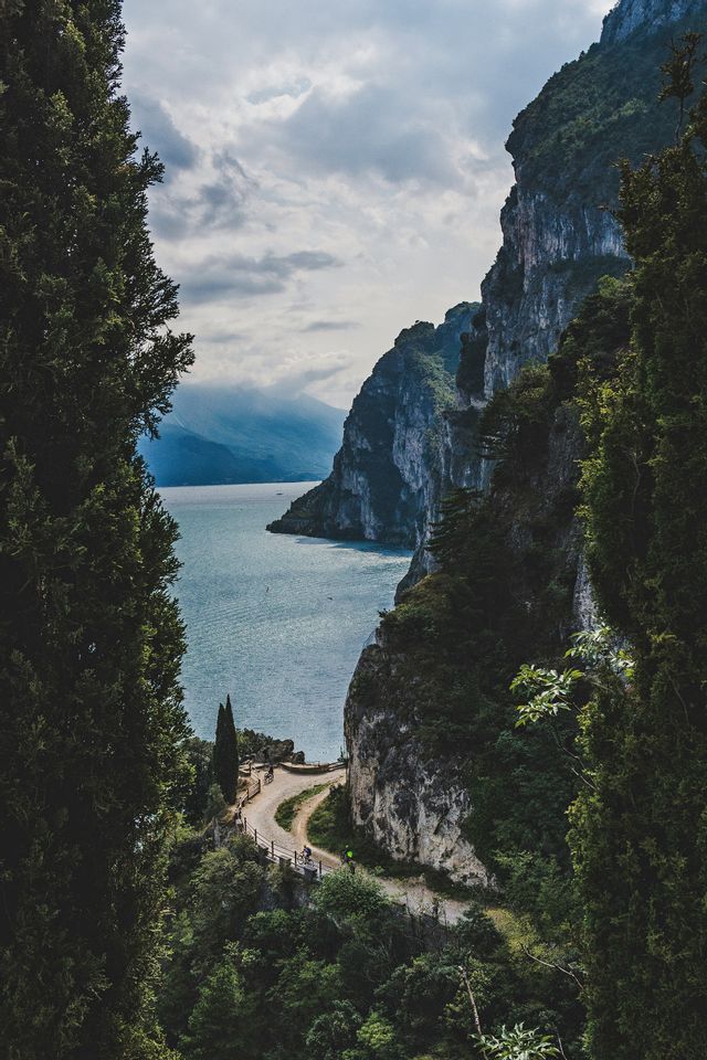 Una vista dall'alto di una strada tortuosa su una parete rocciosa che si affaccia su un grande lago, con ciclisti sul sentiero sottostante.