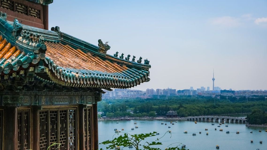 Un tetto tradizionale di pagoda cinese con tegole arancioni e verdi si affaccia su un lago con barche e un paesaggio urbano sullo sfondo.