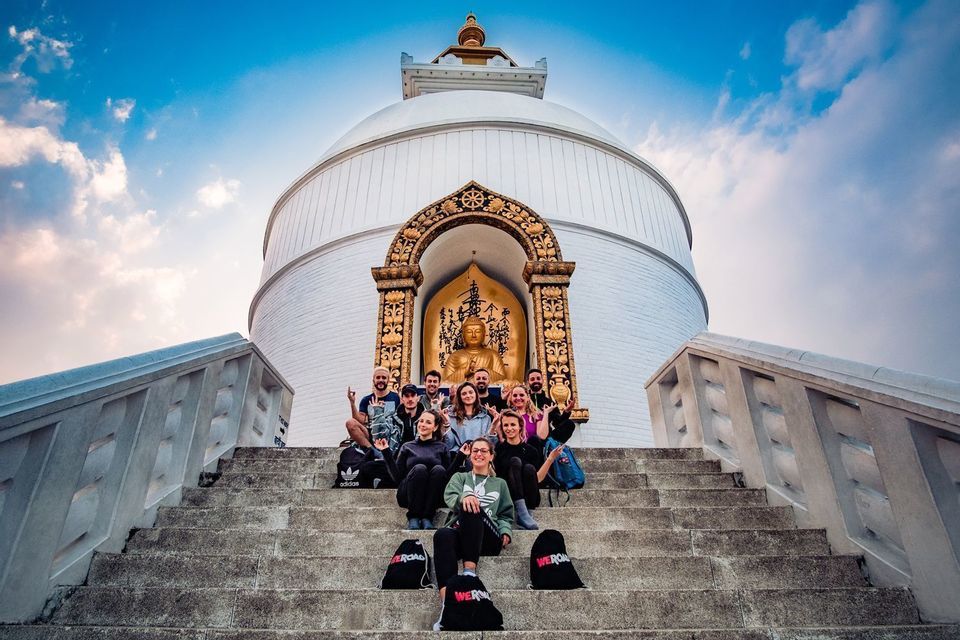 Eine WeRoad-Gruppe posiert für ein Foto auf den Stufen einer weißen Stupa mit goldener Buddha-Statue unter blauem Himmel.