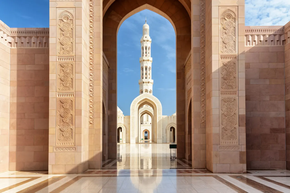 L'alto minareto di una moschea è incorniciato da un grande arco in pietra, con il suo riflesso visibile sul pavimento lucido del cortile.
