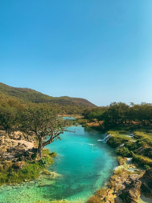 Un fiume limpido e turchese serpeggia attraverso una valle verde e lussureggiante con alberi e colline sotto un cielo azzurro brillante.