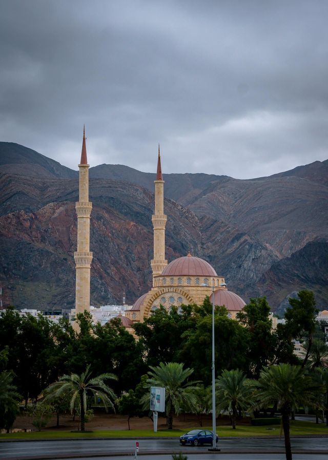 Una moschea con due alti minareti e cupole rosse si staglia contro lo sfondo di montagne rocciose sotto un cielo nuvoloso.