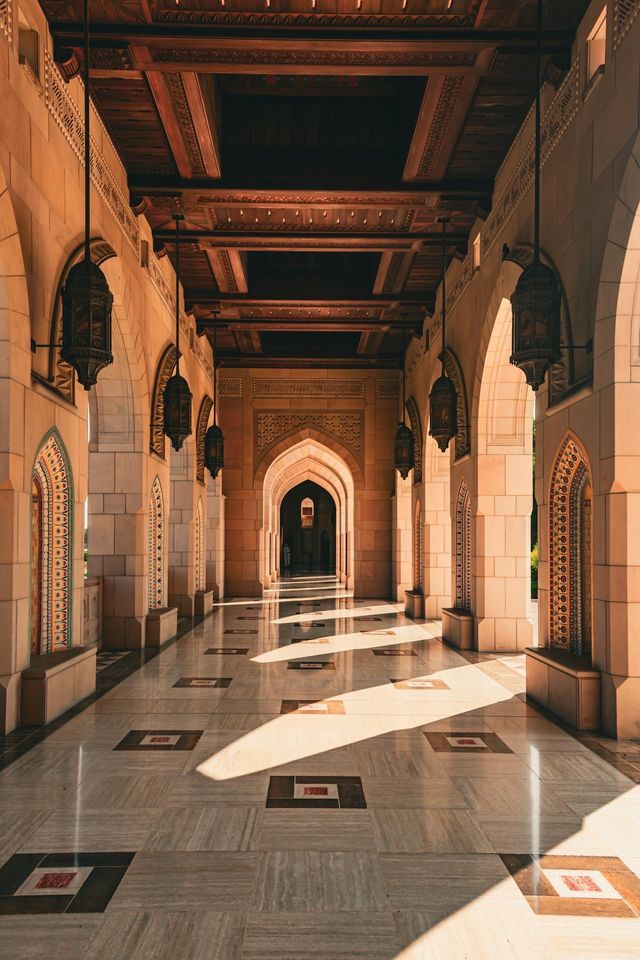 Un corridoio vuoto, illuminato dal sole, con archi in pietra, lanterne pendenti e un pavimento in marmo lucido dai giochi di luce.