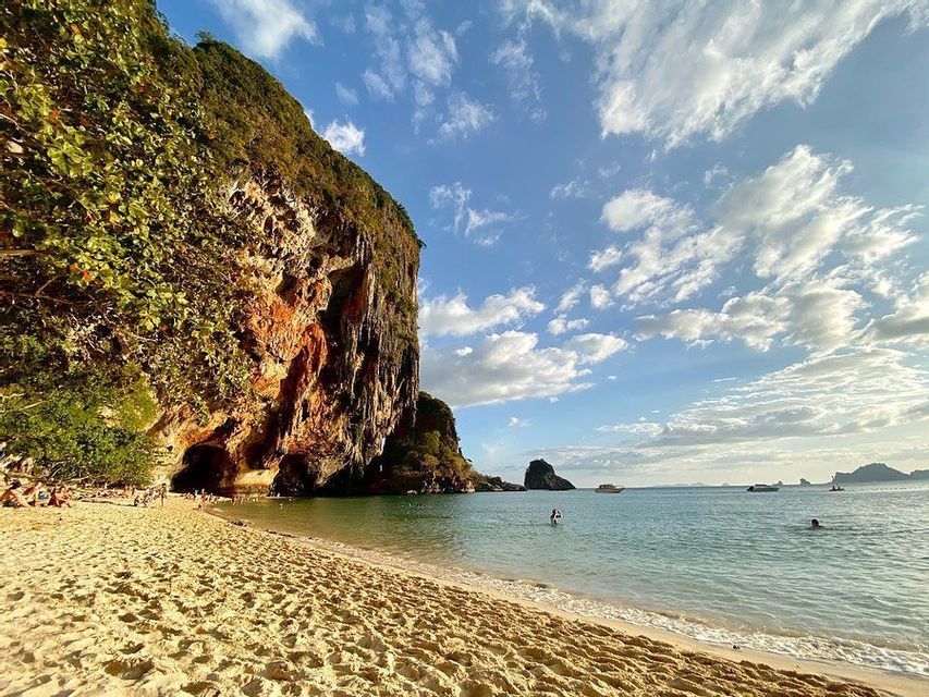 Eine große Felsklippe mit einer Höhle grenzt an einen Sandstrand, an dem Menschen unter einem teilweise bewölkten Himmel schwimmen und entspannen.