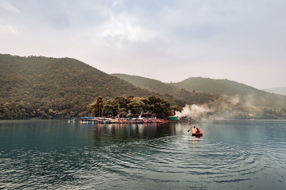 Dos personas reman en un pequeño bote en un lago hacia una pequeña isla, con grandes colinas arboladas al fondo bajo un cielo nublado.