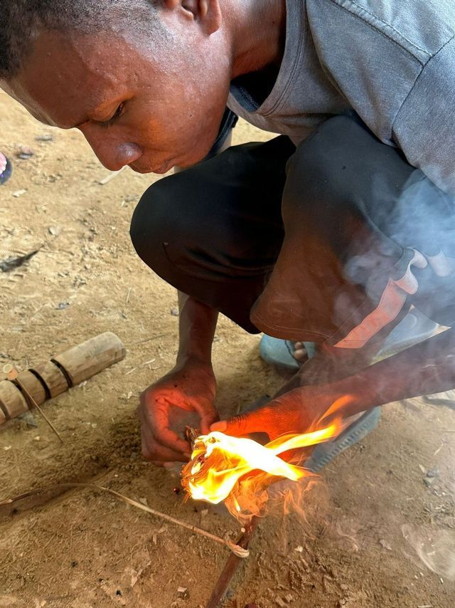 Un uomo si accovaccia a terra e accende con cura un fuoco a mano, soffiando sull'esca fiammeggiante.