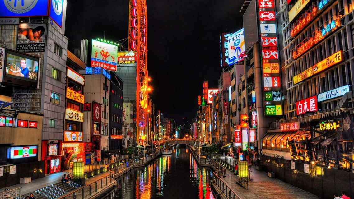 Un canale cittadino di notte fiancheggiato da edifici con insegne al neon giapponesi colorate che si riflettono sull'acqua.