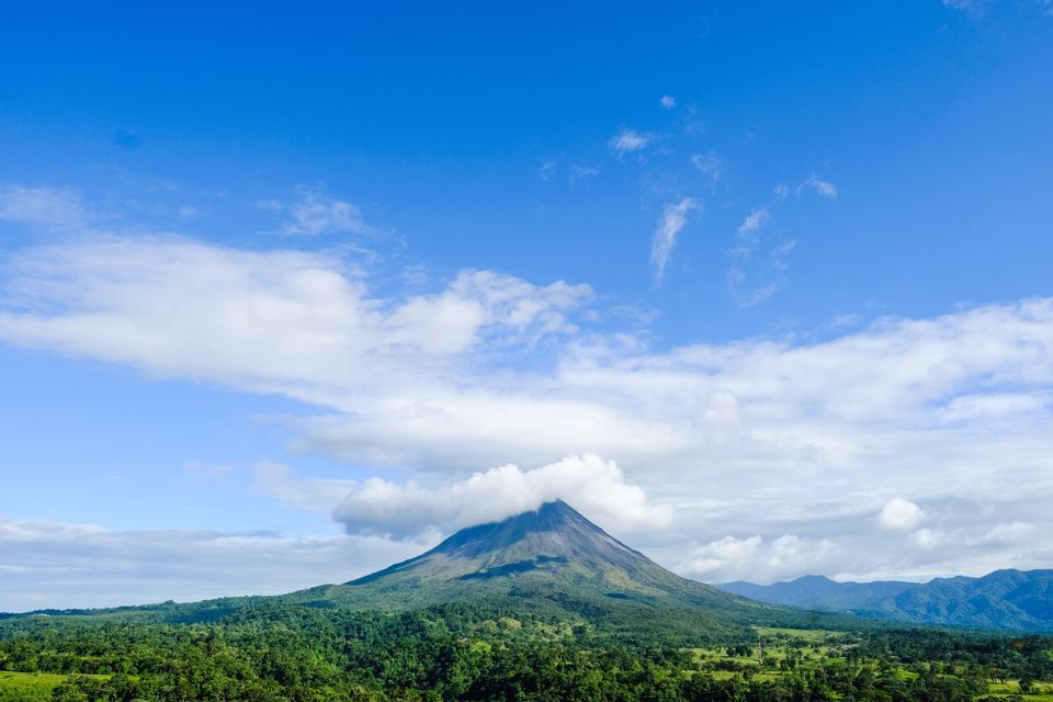 Un grande vulcano con la cima avvolta dalle nuvole, che si erge sopra una fitta foresta verde sotto un cielo azzurro brillante.