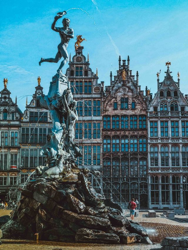 Una statua di bronzo in cima a una grande fontana spruzza acqua in una piazza storica di fronte a edifici ornati.